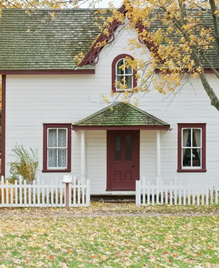 White clapboard house with a red door and a picket fence under autumn maples